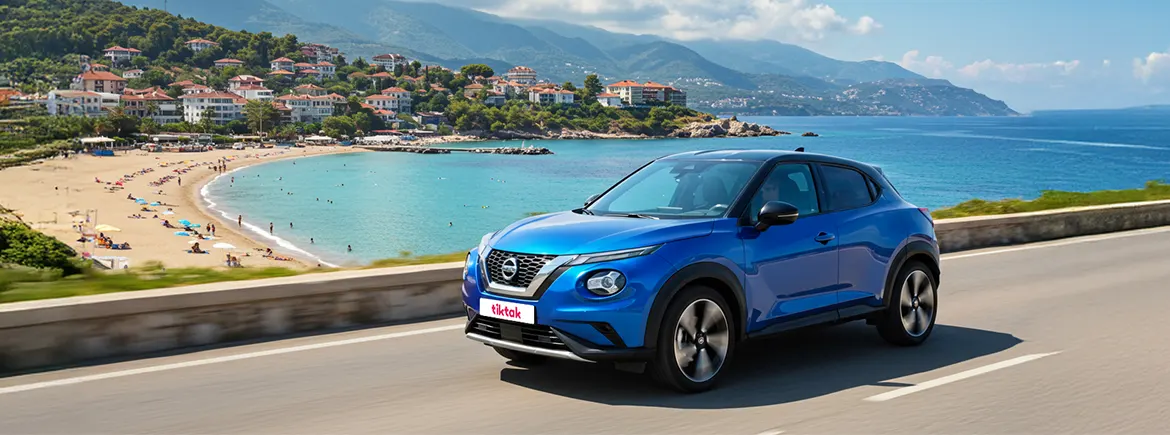Mavi bir Nissan SUV araba, sahil yolunda ilerlerken, arka planda plaj, deniz ve dağlık bir kasaba manzarası ile güneşli bir gün görüntüsü.