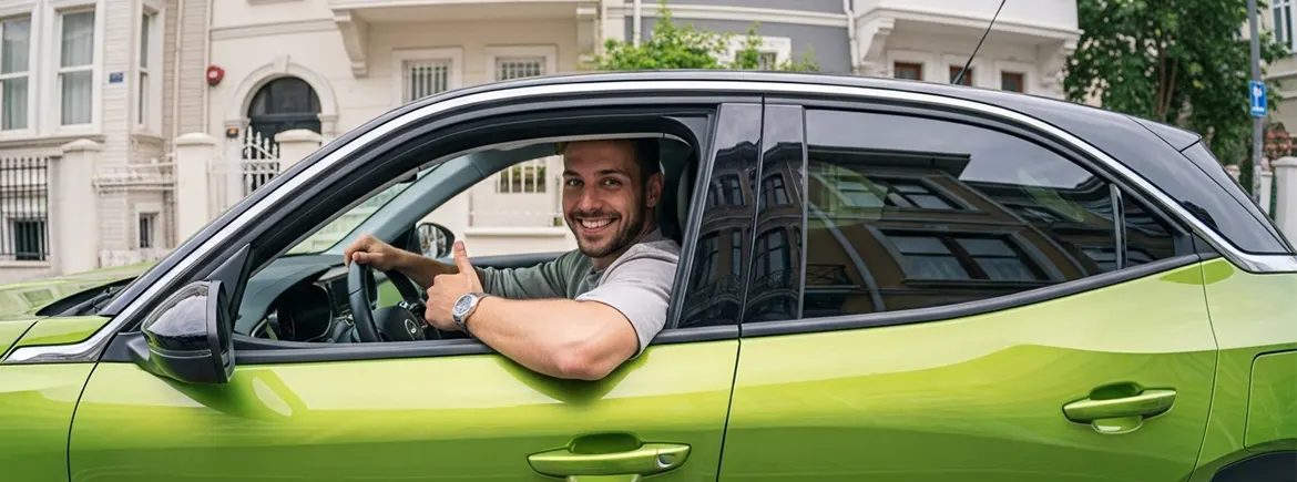 Gülümseyen bir sürücü, yeşil bir SUV'un içinde direksiyon başında oturuyor ve başparmağıyla onay işareti yapıyor. Arka planda şık bir mahalle manzarası var.