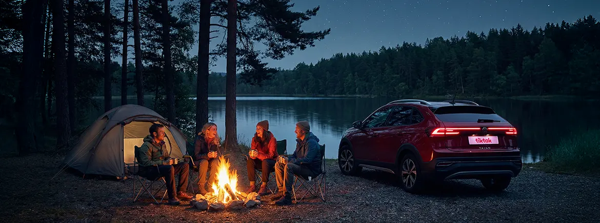 Yıldızlı bir gecede göl kenarında kamp ateşi başında sohbet eden arkadaşlar ve hafta sonu kaçamağı için günlük araç kiralama ile gidilen kırmızı Volkswagen Taigo.